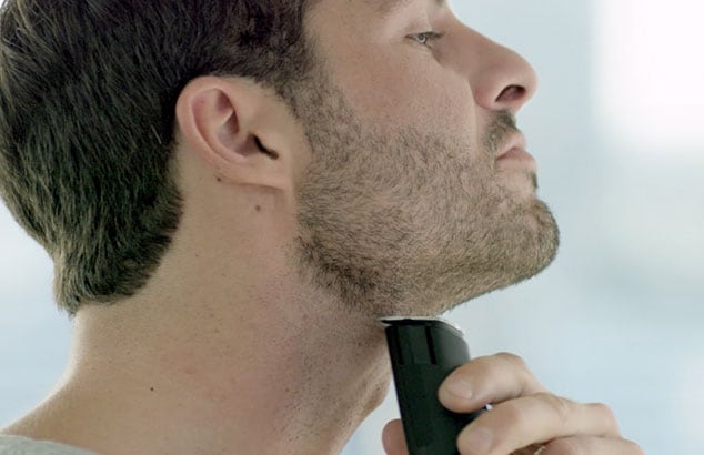 Boční profil muže s krátkými vousy, který si holí krk elektrickým holicím strojkem.