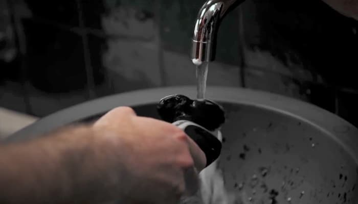Detailní záběr na muže, který čistí elektrický holicí strojek pod tekoucí vodou.
